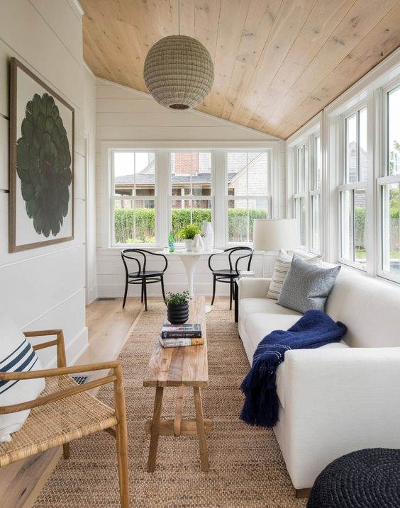 a neutral modern sunroom with a white sofa, some pillows, a white table and black chairs, a wooden coffee table and a woven chair