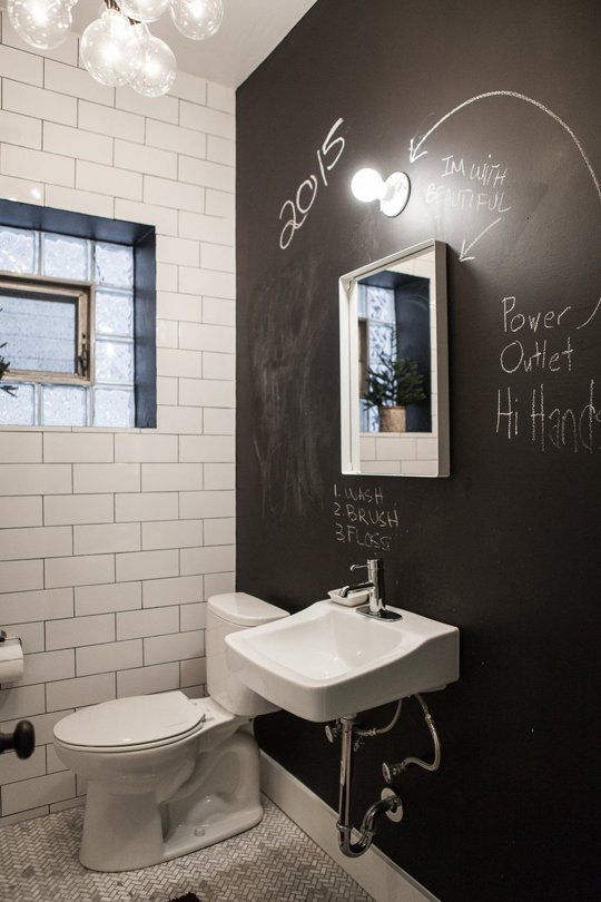 a powder room with a chalkboard accent wall, white tiles, a wall mounted sink,a  toilet and a bubble chandelier