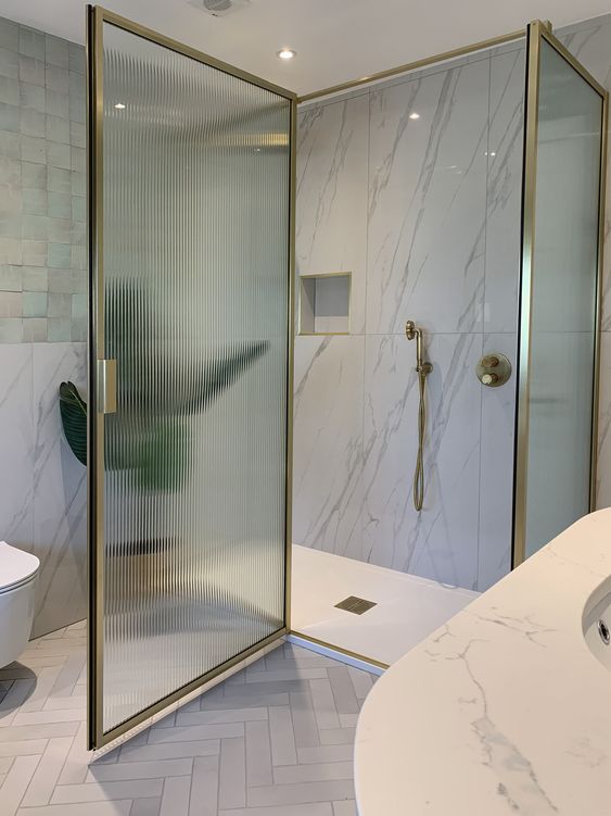 a refined bathroom with white marble tiles, a shower space with ribbed glass doors in gold frames and gold fixtures is wow