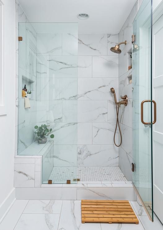 a refined shower space with white marble and penny tiles, glass doors and brass fixtures plus greenery in a vase