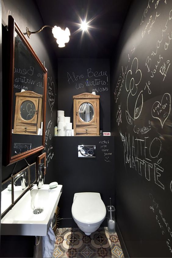 a small and cool powder room with black chalkboard walls, a wall mounted sink, a mirror in a frame and a tiled floor