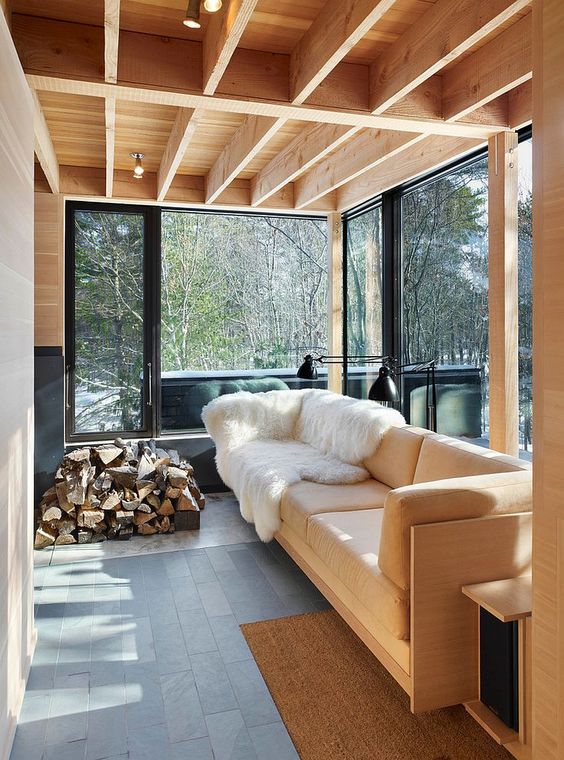 a small modern sunroom with a light-colored sofa and a side table, firewood stacked and a couple of black lamps