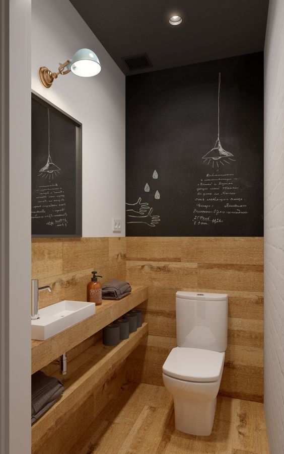 a small powder room with a chalkboard wall, timber paneling, a built in sink, a mirror and a sconce