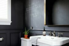 a stylish modern powder room with chalkboard walls, a white floor, a pedestal sink, a white toilet, a mirror in a brass frame and a pendant lamp