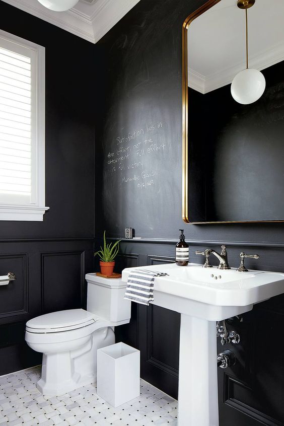 a stylish modern powder room with chalkboard walls, a white floor, a pedestal sink, a white toilet, a mirror in a brass frame and a pendant lamp