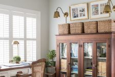 a vintage farmhouse home office with a white desk, a rattan chair and a storage unit, baskets and brass scones
