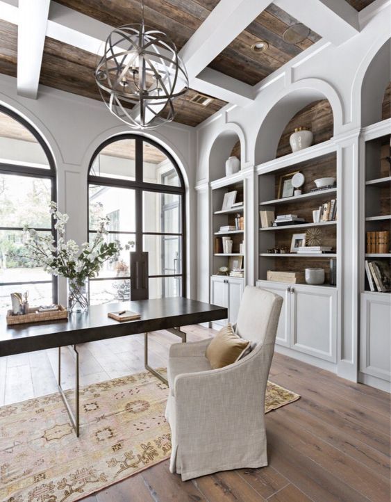 a vintage farmhouse home office with white walls and storage units, a sphere chandelier, a modern desk and an upholstered chair