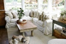 a welcoming farmhouse sunroom with a white L-shaped sofa, some vintage tables, baskets, neutral textiles and potted greenery