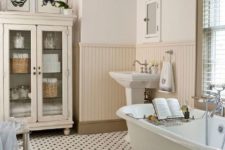 a welcoming neutral bathroom with penny tiles on the floor, sady beadboard, a clawfoot tub and a glass armoire for storage