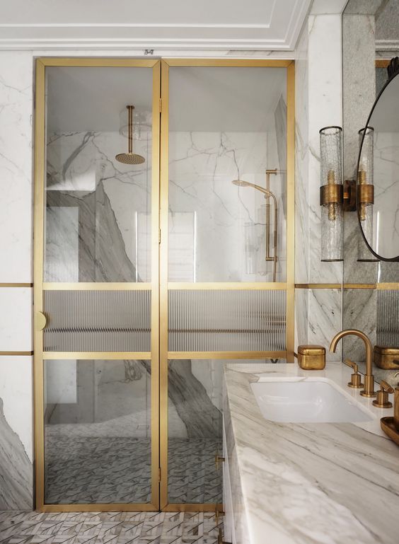 an exquisite bathroom clad with white marble tiles and a vanity, a shower space with glass and gold doors and gold fixtures