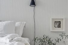 a Scandinavian bedroom finished off with a grey and white striped wall, a bed and a nightstand, some greenery and art is welcoming