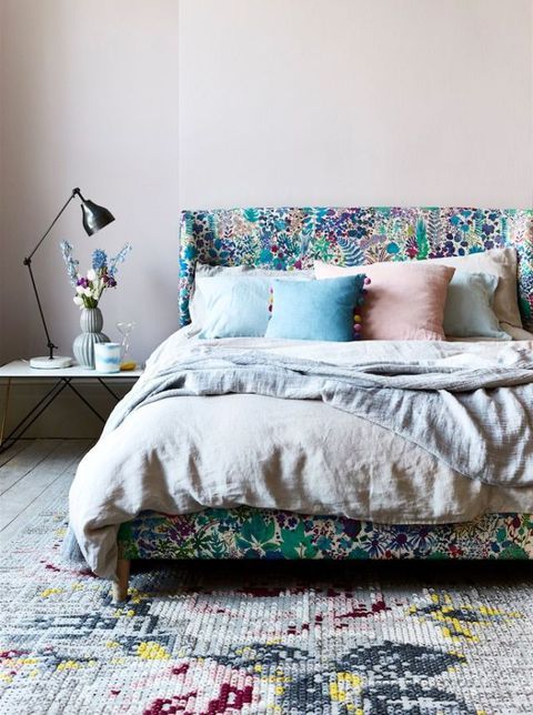 a bright bedroom with a bold upholstered bed and pastel bedding, a floral rug, a nightstand with a black table lamp
