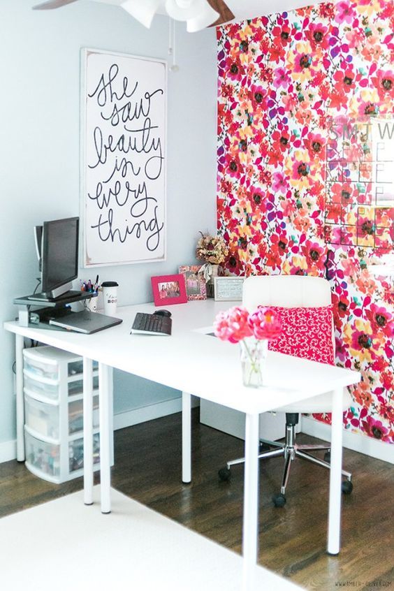 a bright home office with a bold floral accent wall, a white corner desk and a chair with a bright pillow, an artwork