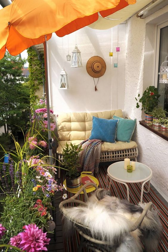 a bright summer balcony with bold textiles, rattan and metal furniture, colorful candle lanterns and an umbrella plus potted blooms