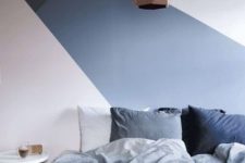 a chic bedroom with a geometric accent wall, a bed with blue and grey bedding, a round nightstand, a faceted pendant lamp
