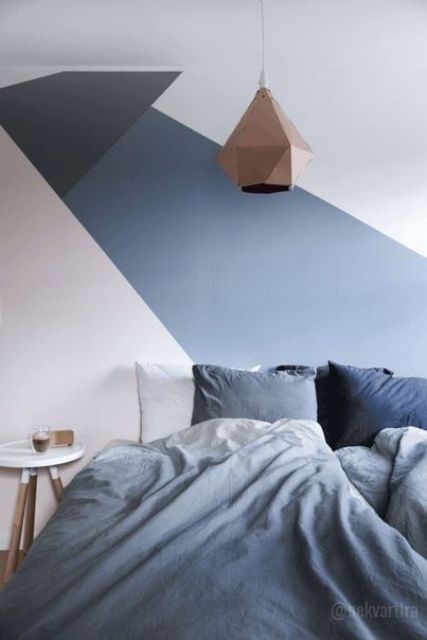 a chic bedroom with a geometric accent wall, a bed with blue and grey bedding, a round nightstand, a faceted pendant lamp