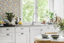 a chic cottage kitchen with white shaker cabinets, a bright floral wallpaper wall, stone countertops and black pendant lamps
