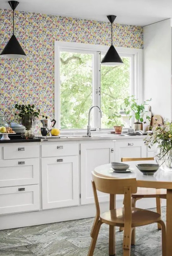 a chic cottage kitchen with white shaker cabinets, a bright floral wallpaper wall, stone countertops and black pendant lamps