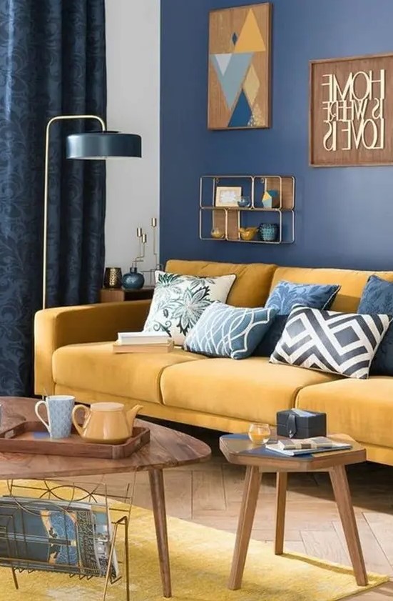 a chic living room with a navy wall, a yellow sofa and a rug, printed pillows, a cool gallery wall and wooden tables