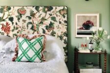 a cool and chic bedroom with green walls, a bed with a floral headboard, neutral bedding and a green bedspread, a nightstand and white blooms