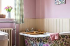 a cottage bathroom with pink walls, neutral paneling, a bright floral clawfoot tub, a printed rug and curtains