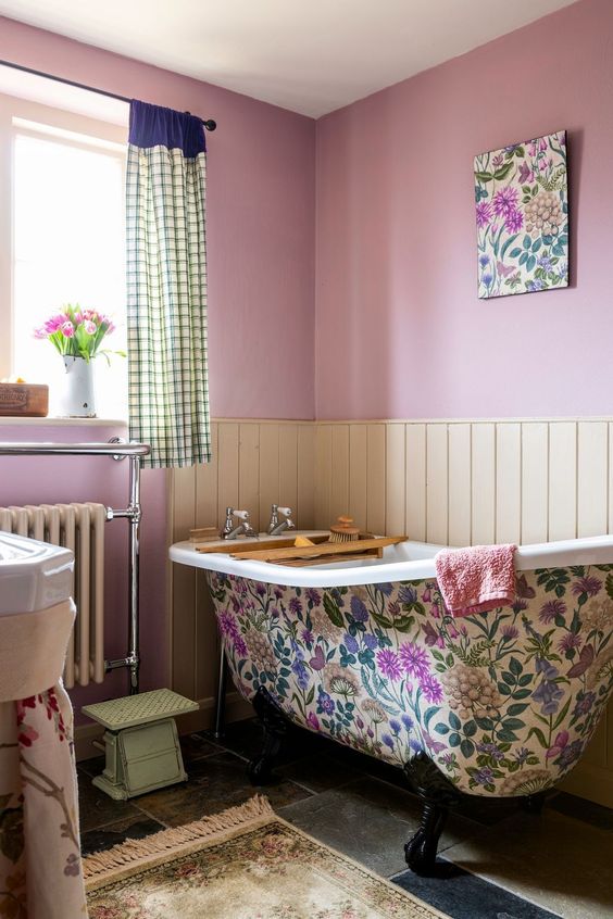 a cottage bathroom with pink walls, neutral paneling, a bright floral clawfoot tub, a printed rug and curtains