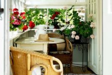 a cozy small sunroom with wicker chairs and a foldable table, with lots of blooms in pots and Roman shades