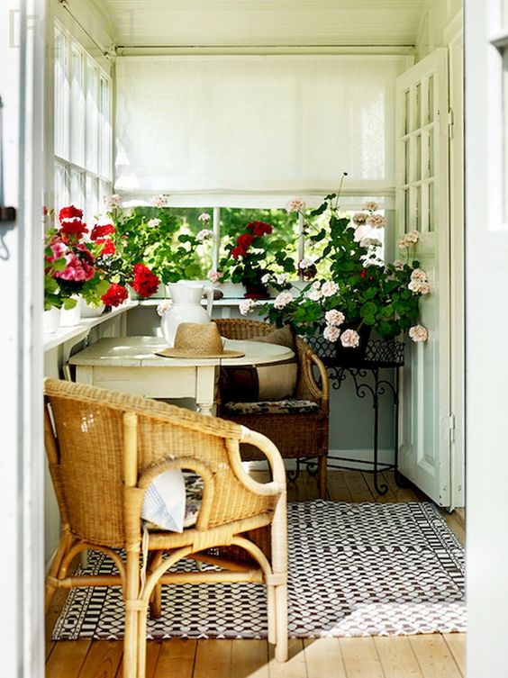 a cozy small sunroom with wicker chairs and a foldable table, with lots of blooms in pots and Roman shades