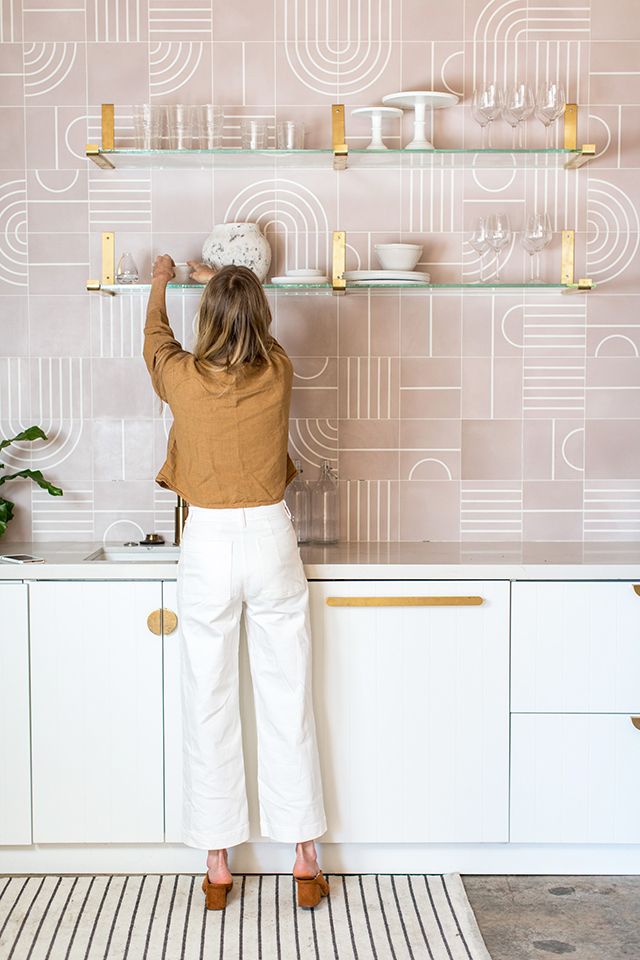 a delicate kitchen with sleek white cabinets, a pink geo tile wall, glass and gold shelves is a very beautiful and unique idea