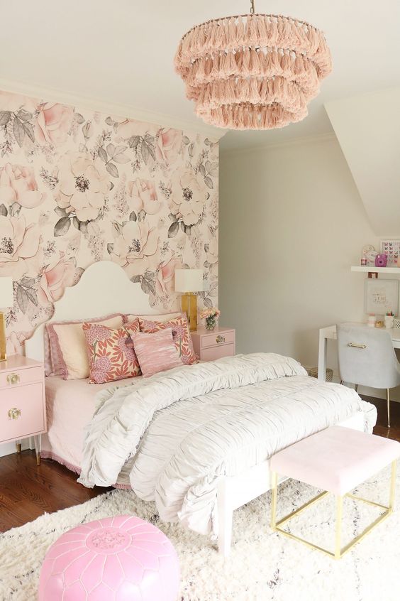 a delicate teen girl bedroom with a blush floral accent wall, a bed with pink bedding, a pink nightstand and pink tassel chandelier