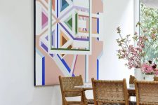 a dining room with a stained table and woven leather chairs, a pink rug, a colorful geometric artwork and blooms and greenery