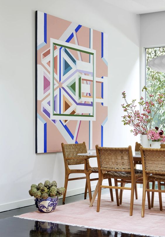 a dining room with a stained table and woven leather chairs, a pink rug, a colorful geometric artwork and blooms and greenery