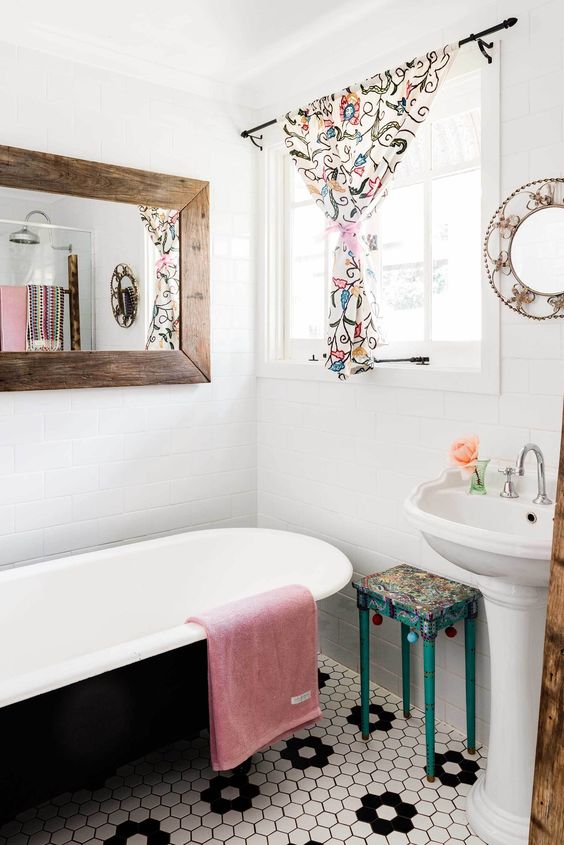 a farmhouse bathroom with a hex tile floor, a black bathtub, a mirror in a stained frame, a bold floral curtain and stool