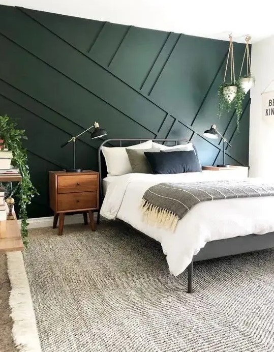 a mid-centiry modern bedroom with a dark green geometric panel wall, a metal bed, wooden furniture and some greenery in pots to enliven it