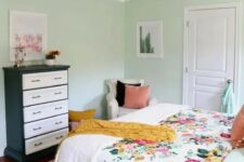 a mint-colored bedroom with a bed and floral bedding, a black and white dresser, a neutral chair and a chandelier