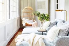 a modern coastal sunroom with striped daybeds, white pillows and blankets, a hanging rattan chair and greenery