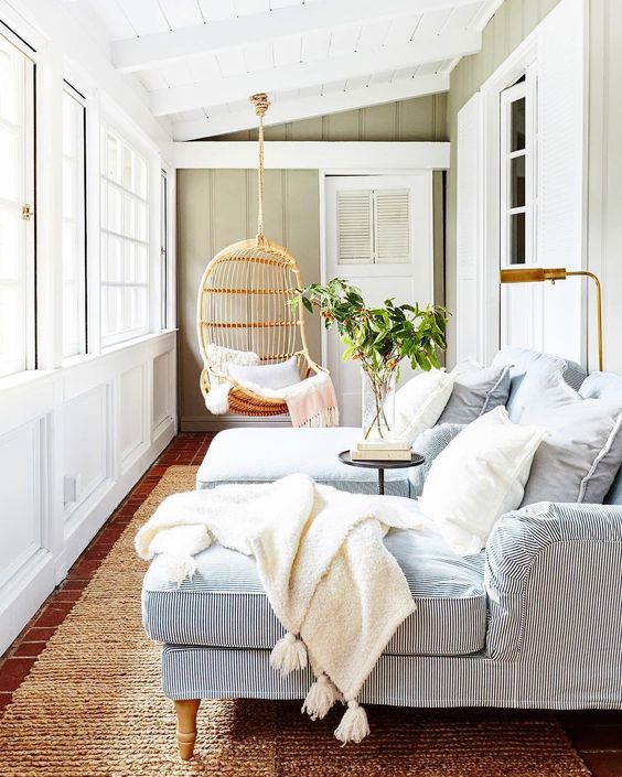 a modern coastal sunroom with striped daybeds, white pillows and blankets, a hanging rattan chair and greenery