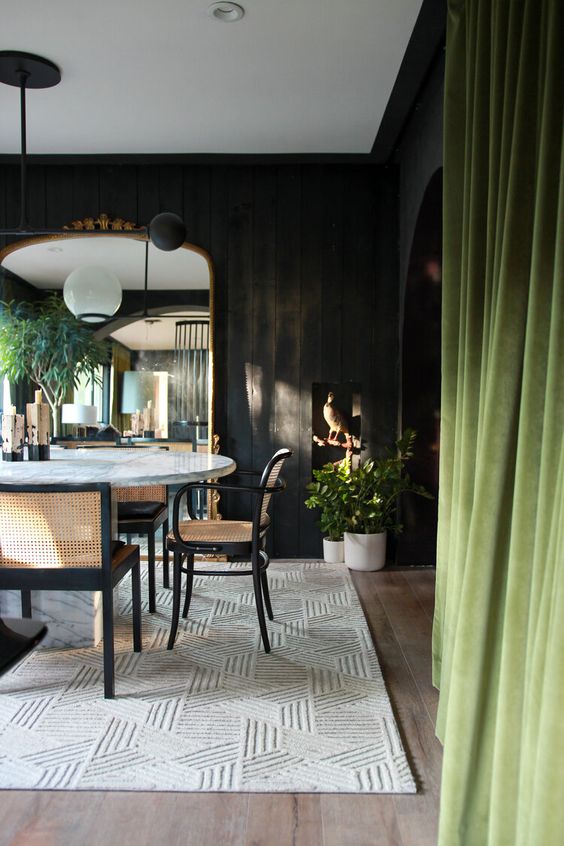 a moody dining room with black walls, a round dining table, cane chairs, mid-century modern lamps and a chic geo printed rug