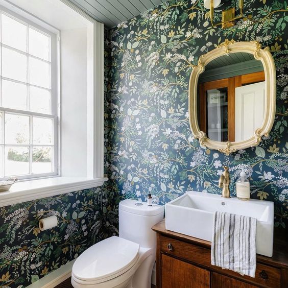 a moody powder room with dark green floral wallpaper, a stained vanity, a mirror in a chic frame and white appliances