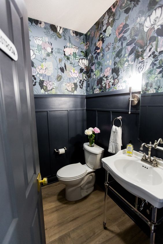a moody powder room with grey floral wallpaper and black panling, a free standing sink and a toilet, some blooms