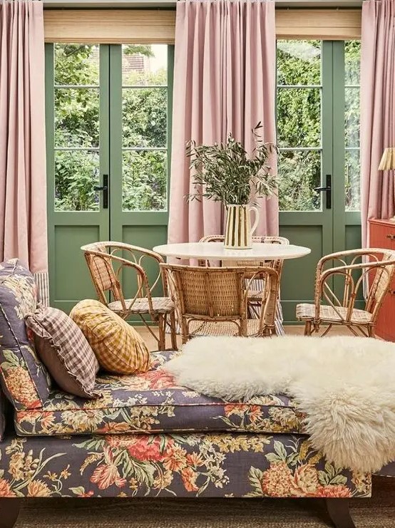 a muted color living room with green walls and doors, pink curtains, a floral daybed and a rattan dining nook