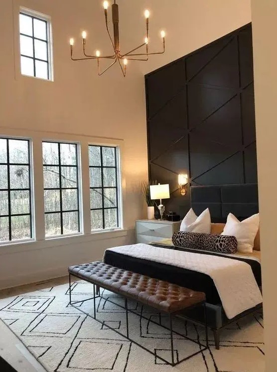 a refined bedroom with a black paneled geometric wall, a black upholstered bed, a leather bench and a catchy chandelier