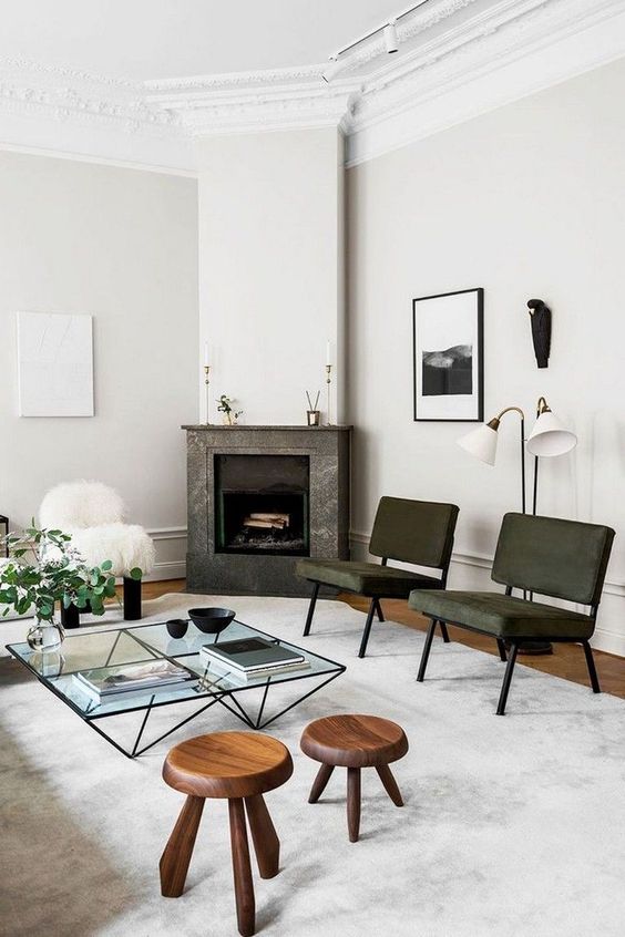 a refined living room with a stone fireplace, a glass coffee table with a geometric base, wooden stools and dark green leather chairs
