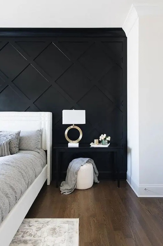 a refined monochromatic bedroom with a black geometric paneled wall, a white bed, a black nightstand, a white pouf and a chic lamp