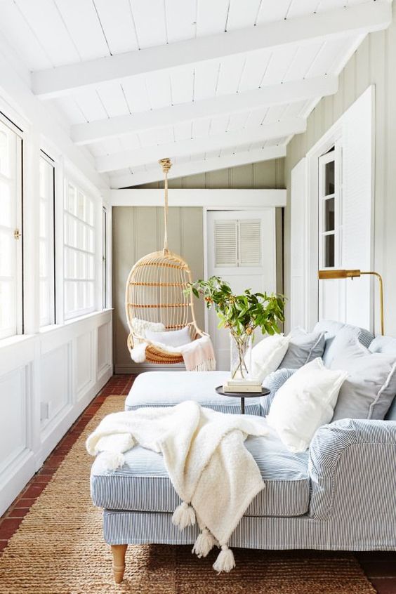 a relaxing small sunroom with striped blue lounge chairs, a lamp, a table and a hanging wicker chair