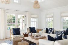 a seaside living room with wooden beams and lamps hanging on them, white seating furniture, neutral chairs and navy pillows, a wooden coffee table and jute poufs plus a striped rug