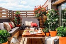a summer balcony with a wooden deck and furniture, potted blooms and greenery and bright pillows plus candle lanterns