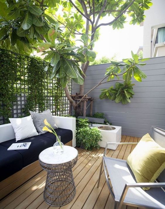 a tiny modern terrace with a wooden deck, a wall with greenery, a built-in sofa, a chair and a fountain plus a small table