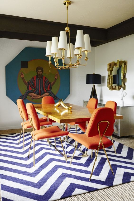 a unique dining space with a creative artwork, a mirror in a gold frame, a navy and white chevron printed rug, orange and gold chairs and a chic stone table