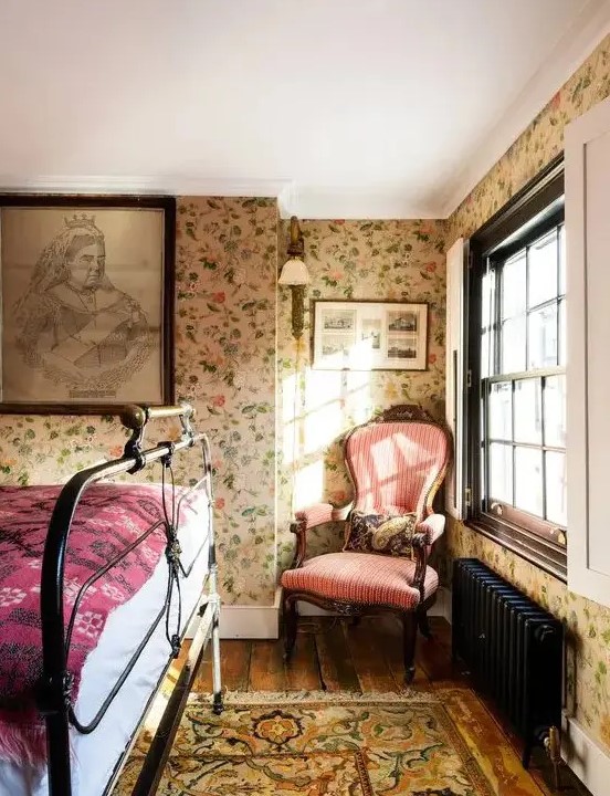 a vintage bedroom with floral wallpaper, a black forged bed, a vintage pink chair and printed textiles